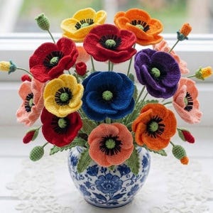 Op de afbeelding: Een boeket van gehaakte klaprozen in levendige tinten rood, geel, oranje, blauw, paars en perzik, elk met een groen hart en zwarte meeldraden. De bloemen zijn geschikt in een ronde, witte keramische vaas met blauwe bloemmotieven.