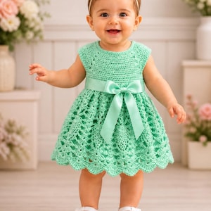 May include: A mint green crochet dress with a matching bow headband and satin ribbon sash. The dress features a scalloped hem and is paired with white Mary Jane shoes. A baby is wearing the dress and smiling.