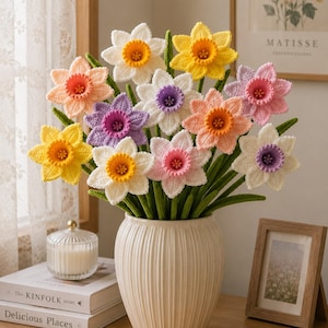 Può includere: Un bouquet di fiori di narciso all'uncinetto in vari colori tra cui bianco, giallo, pesca e lavanda, ognuno con una tromba centrale. I fiori hanno steli verdi e sono disposti in un vaso di ceramica a coste color bianco sporco.