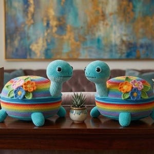 May include: Two handmade crochet turtle-shaped decorative items. Each turtle has a rainbow shell, blue head, and legs. The shells are adorned with colorful flower appliques. They are placed on a dark wooden table.