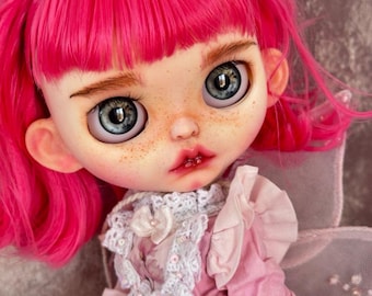 May include: A doll with bright pink hair, grey eyes, and freckles, wearing a pink dress with a white crocheted collar and a floral headpiece. The doll features a pale complexion, rosy cheeks, and pink lips, with sheer pink wings visible on the back.