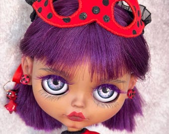 May include: A doll with vibrant purple hair wearing a red mask with black polka dots. The doll features large, detailed light-colored eyes with long lashes, red lips, and matching red and black polka dot earrings. A red and black polka dot top is visible.