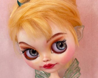 May include: A doll with a detailed face, blonde hair, and blue eyes. The doll has a green top and translucent green wings. A green bow is in the doll's hair. The doll's face has rosy cheeks and red lips.