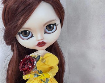 May include: A doll with long auburn hair, large brown eyes, and dark red lipstick. The doll wears a bright yellow off-the-shoulder dress with a small gold embellishment at the center. It holds a dark red rose with silver glitter accents.