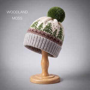 May include: A knit beanie with a folded brim, featuring a pattern of green pine trees and brown geometric shapes on a cream background. The hat is topped with a large, textured green pom-pom. The text WOODLAND MOSS is visible.
