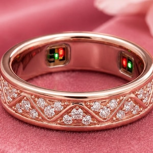 May include: A rose gold smart ring featuring a continuous zigzag pattern embedded with small clear stones. The interior of the band displays two small rectangular sensor modules with visible green and red light indicators.