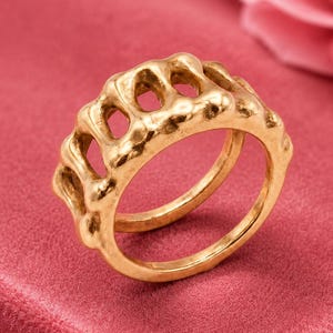 May include: A gold-toned ring featuring a continuous, open-work band with a series of rounded, interconnected vertical pillars. The surface of the metal has a textured, organic finish, creating a repeating pattern of small, oval-shaped cutouts around the entire circumference of the band.