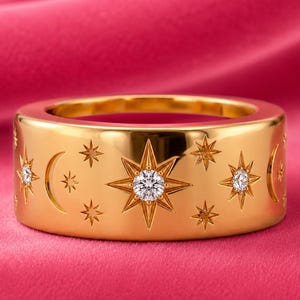May include: A wide gold band ring featuring an engraved celestial pattern of stars and crescent moons. The design includes three small, clear, round-cut stones set into the centers of the largest star motifs, creating a polished, reflective finish.