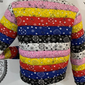 May include: A multi-colored puffer jacket made from patchwork bandana fabric. The jacket has a horizontal striped pattern with alternating colors of pink, yellow, red, blue, black, and white. The jacket has a zipper closure and a drawstring hood. The label on the jacket reads "Lemonade Millionaires".