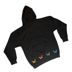 May include: Black hooded sweatshirt with four embroidered butterflies in rainbow colors on the bottom hem.