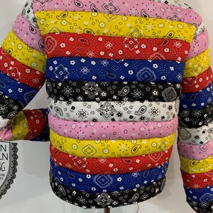 May include: A multi-colored puffer jacket with a horizontal striped pattern. The jacket is made of different bandana fabrics in shades of pink, yellow, red, blue, and black. The jacket has a drawstring at the bottom and a label that reads "Lemonade Millionaires".