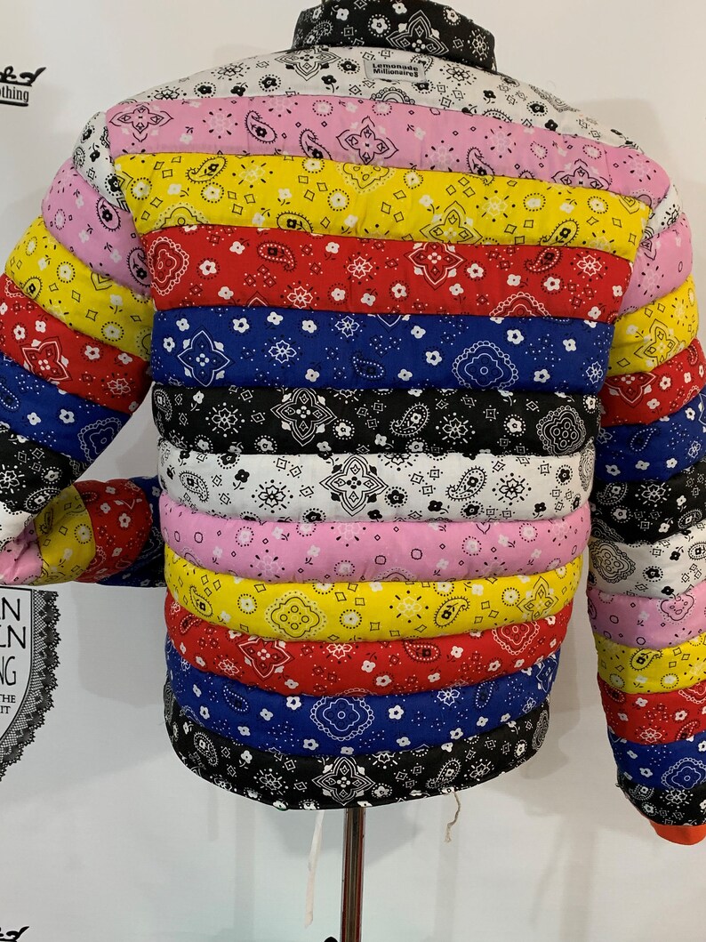 May include: A multi-colored puffer jacket with a horizontal striped pattern. The jacket is made of different bandana fabrics in shades of pink, yellow, red, blue, and black. The jacket has a drawstring at the bottom and a label that reads "Lemonade Millionaires".