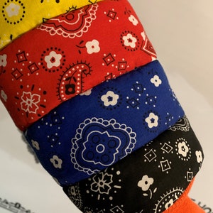 May include: A stack of four bandanas in different colors, yellow, red, blue, and black, with a white paisley pattern. The bandanas are folded and stacked on top of each other.
