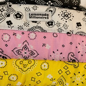 May include: A stack of five bandanas in different colors and patterns. The top bandana is black and white, the second is white with black designs, the third is pink with white designs, the fourth is yellow with black designs, and the bottom bandana is red with black designs. The second bandana has a label that reads "Lemonade Millionaire".