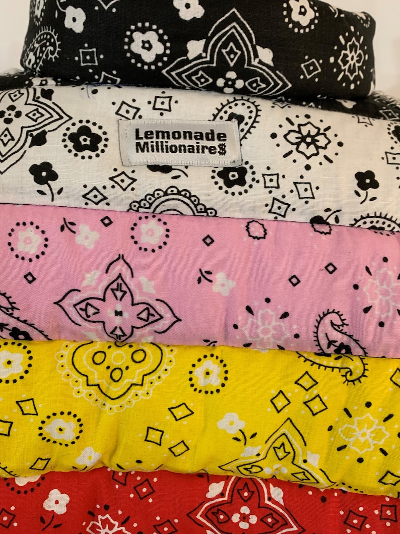 May include: A stack of five bandanas in different colors and patterns. The top bandana is black and white, the second is white with black designs, the third is pink with white designs, the fourth is yellow with black designs, and the bottom bandana is red with black designs. The second bandana has a label that reads "Lemonade Millionaire".