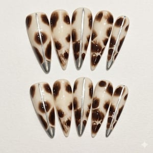 Peut inclure: Un ensemble de dix faux ongles en forme d'amande. Les ongles présentent une base crème avec des taches marron, une bande argentée au centre et des pointes argentées. Le motif ressemble à un motif de peau de vache.