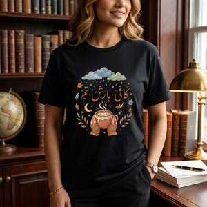 May include: A black t-shirt featuring a graphic illustration of hands holding a steaming mug of coffee. Above the mug are Arabic script, a crescent moon, falling leaves, and rain clouds. The design incorporates warm autumn colours and soft blue tones.