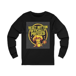 Puede incluir: Una camiseta negra de manga larga con un estampado gráfico de un brócoli amarillo con ojos en forma de X y expresión triste dentro de un círculo amarillo. El texto dice RESET BUTTON OFFICIALLY PRESSED con un icono de cero a la derecha.