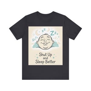 May include: A black t-shirt featuring a graphic of a sleeping face with a piece of tape over the mouth, surrounded by blue Zzz symbols, stars, and a crescent moon. Below the illustration, the text reads Shut Up and Sleep Better in black.