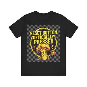 Puede incluir: Una camiseta negra de manga corta con un estampado gráfico de un brócoli triste con ojos en forma de X dentro de un círculo amarillo. El texto dice RESET BUTTON OFFICIALLY PRESSED, acompañado de pequeños iconos de rayos y un símbolo cero.