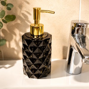 Peut inclure: Un distributeur de savon noir avec une surface géométrique à facettes et des éclats dorés. Le distributeur est doté d'un mécanisme de pompe doré brillant sur le dessus, conçu pour distribuer du savon liquide dans une salle de bain ou une cuisine.