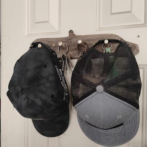 May include: A wooden key and hat holder with two baseball caps suspended from it. One cap is black with a camouflage design, and the other is grey and black with a mesh back. Keys are hanging from the holder.