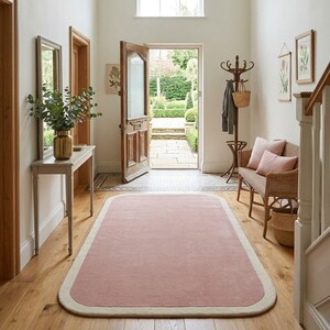 Puede incluir: Una alfombra rectangular con esquinas redondeadas, que presenta un centro de color rosa empolvado sólido y un borde grueso de color crema. La alfombra está colocada sobre un suelo de madera clara en un vestíbulo luminoso.