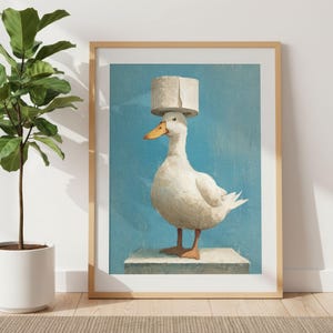 Può includere: Stampa artistica incorniciata raffigurante un'anatra bianca con un rotolo di carta igienica come cappello. L'anatra è su una piccola piattaforma contro uno sfondo blu. L'opera è presentata in una cornice di legno marrone chiaro.