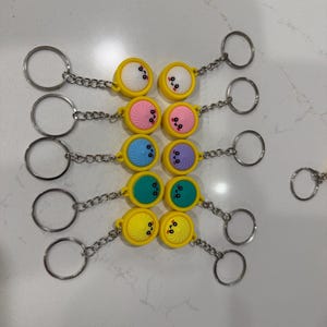 May include: A collection of keychains featuring colorful, round charms with smiling faces. The charms are yellow, pink, blue, green, and purple, and are attached to silver key rings via a chain.