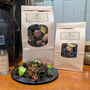 May include: Apothecary spray bottle and two brown paper bags with a clear window, filled with dried botanicals. A small black plate holds a collection of dried plants and green accents. The products are displayed on a wooden surface.
