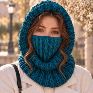 Pode incluir: Uma balaclava de malha azul-petróleo com capuz texturizado e seção de aquecedor de pescoço canelado. A peça é feita com um padrão de tricô grosso ao redor da abertura do rosto e um design de ponto canelado vertical no nariz e pescoço.
