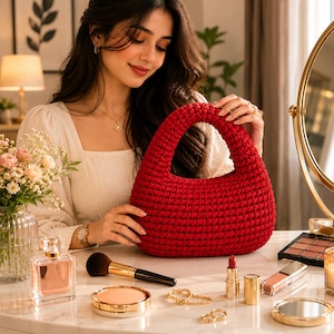May include: A deep red, crescent-shaped handbag featuring a dense, textured crochet knit pattern with a rounded integrated handle. The bag has a structured, curved silhouette and a solid, uniform color throughout its entire exterior surface.