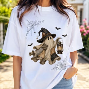 Peut inclure: Un t-shirt blanc orné d'un motif représentant un fantôme portant un chapeau de sorcière en patchwork et tenant une lanterne allumée. Le dessin inclut des toiles d'araignée noires dans les coins et des chauves-souris volant autour du fantôme.