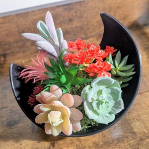 May include: A black, crescent-shaped planter filled with a variety of artificial succulents. The arrangement includes light green, red, and pink faux plants, set against a bed of green moss. The planter sits on a wooden surface.