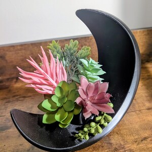 May include: A black, crescent moon-shaped planter filled with artificial succulents in shades of pink, green, and burgundy. The planter is displayed on a wooden surface.