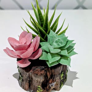 May include: A decorative succulent arrangement featuring a pink flower, a green succulent, and a tall green plant. The plants are set in a brown, wood-like pot with moss accents, creating a natural aesthetic.