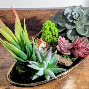 Peut inclure: Composition décorative de succulentes dans un récipient ovale à bordure dorée. L'arrangement comprend diverses succulentes artificielles vertes, roses et rouges, une maison miniature et un petit arbre vert. Le récipient est posé sur une surface en bois.