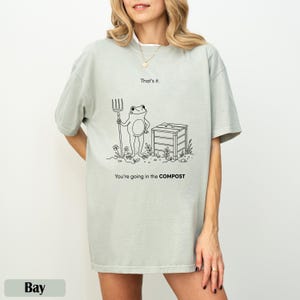Könnte beinhalten: Hellgrünes T-Shirt mit einer Schwarz-Weiß-Illustration eines Frosches, der eine Mistgabel neben einem Kompostbehälter hält. Der Text auf dem Shirt lautet "That's it." und "You're going in the COMPOST."