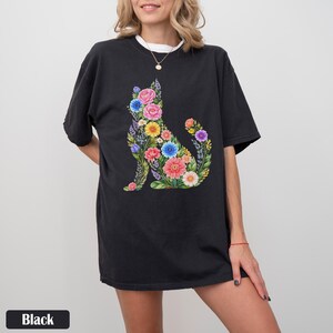 May include: Black t-shirt featuring a floral cat design. The cat silhouette is filled with colorful flowers, including pink roses, blue cornflowers, and yellow daisies. The shirt has a relaxed fit and a white undershirt is visible.