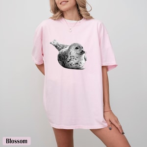 May include: A light pink t-shirt featuring a black and white graphic of a seal. The word "Blossom" is printed at the bottom of the image. The shirt has short sleeves and a relaxed fit.