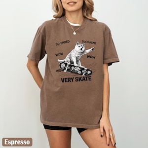 May include: Espresso-colored t-shirt featuring a graphic of a dog riding a skateboard. The dog is white and gray, with the words "SO SHRED," "SUCH MEME," and "VERY SKATE" printed around the image. The shirt has short sleeves.