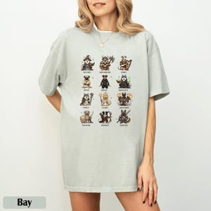 May include: A light gray t-shirt featuring a grid of illustrated dogs dressed as fantasy characters, including a wizard, barbarian, and bard. The shirt has a relaxed fit and is made of a soft material. The word "Bay" is at the bottom.
