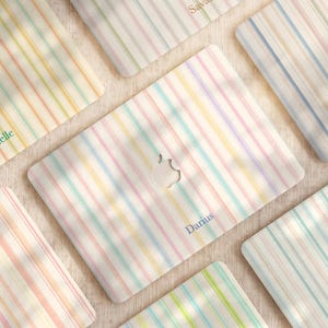 May include: Multiple laptop skins featuring a vertical striped pattern in pastel shades of pink, yellow, blue, and green. Each skin has a central cutout for an Apple logo and is personalized with a name such as Ramona, Savannah, Brielle, Darius, Cedric, or Hector.