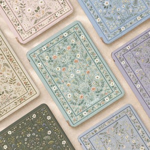 Pode incluir: Uma coleção de capas retangulares para tablets com vários fundos em tons pastel e suaves, cada uma decorada com um padrão botânico detalhado de flores silvestres e uma moldura central, com nomes personalizados impressos na parte inferior.