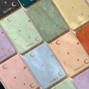 May include: A collection of rectangular, velvet-textured notebooks in various pastel and dark colors, each featuring a gold-foiled celestial pattern of moons and stars. Several notebooks are personalized with names like Bonnie, Stella, Adeline, Clover, Brynn, Eleanor, and Dorothy.
