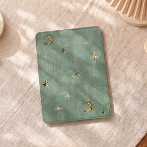 May include: A sage green rectangular mat featuring a celestial design with gold moons and stars. The mat is placed on a light-coloured, textured surface. The design is reminiscent of a night sky.