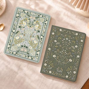 Puede incluir: Dos cuadernos rectangulares con motivos florales. El cuaderno de la izquierda es de color verde salvia claro con el nombre Sophia impreso en la parte inferior. El de la derecha es verde oliva oscuro con el nombre Trista. Ambos muestran ilustraciones botánicas de flores blancas, amarillas y azules.