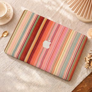 Può includere: Una skin per laptop con un motivo a righe verticali in varie tonalità di rosa, arancione, rosso, giallo e verde acqua. Il design copre la superficie rettangolare del laptop, che presenta un logo Apple argentato centrato sul coperchio.