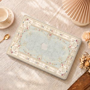 Op de afbeelding: Een laptophoes met een lichtblauw en crèmekleurig bloemmotief, dat lijkt op een vintage tapijt. De hoes heeft een Apple-logo-uitsparing en een decoratieve rand met roze en gouden accenten. De hoes is ontworpen voor een laptop.