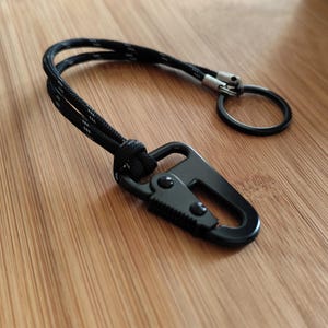 Op de afbeelding: Zwarte karabijnhaak sleutelhanger met een zwart-wit touw koord en een splitring. De karabijnhaak heeft een veergeladen poort en is gemaakt van metaal. De sleutelhanger ligt op een houten oppervlak.